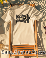 Beige t-shirt with 'Desert Club' design on a wooden chair with palm leaves and sunglasses.